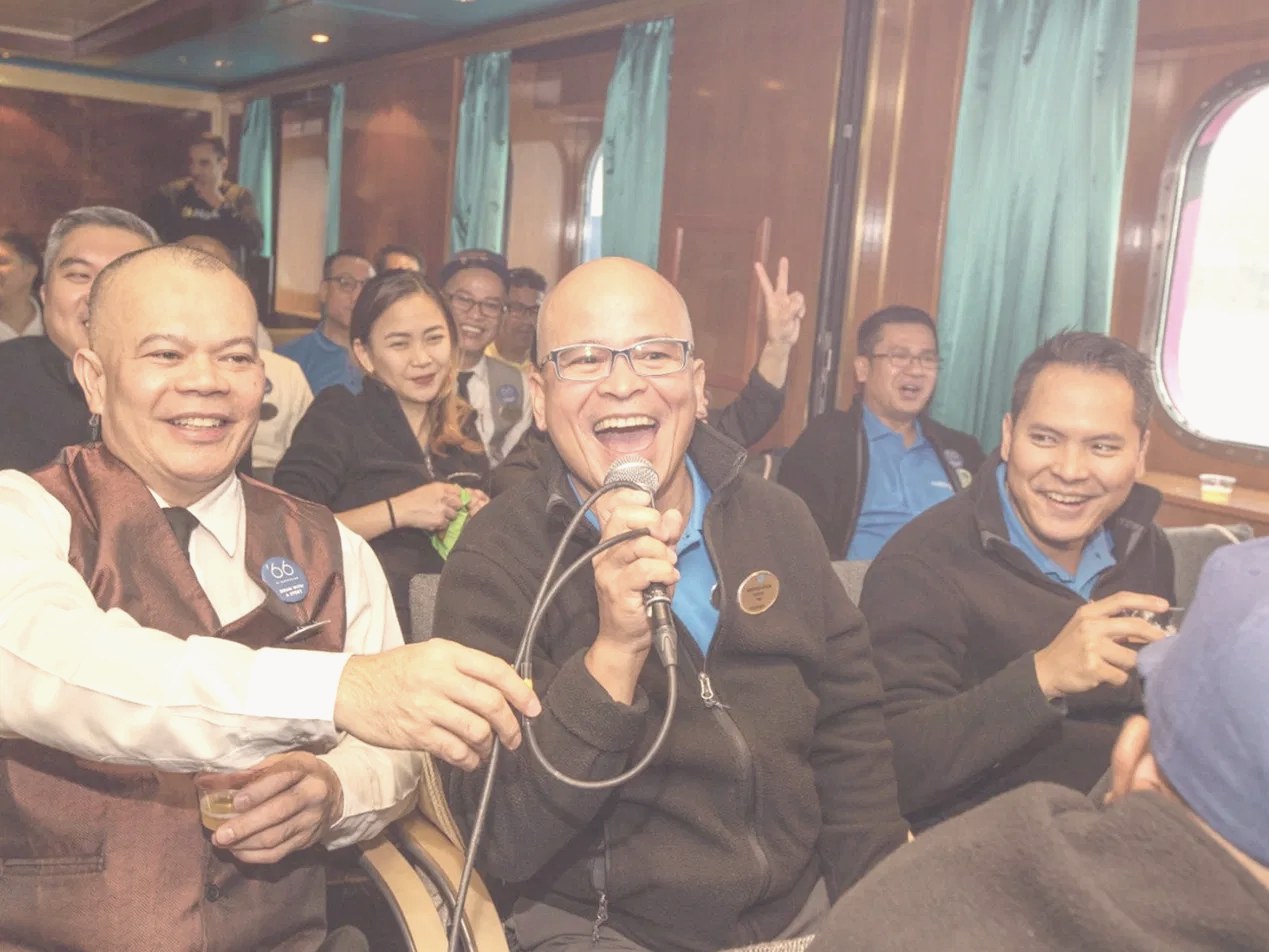 Guests laughing together as a host sings into a microphone at an onboard event